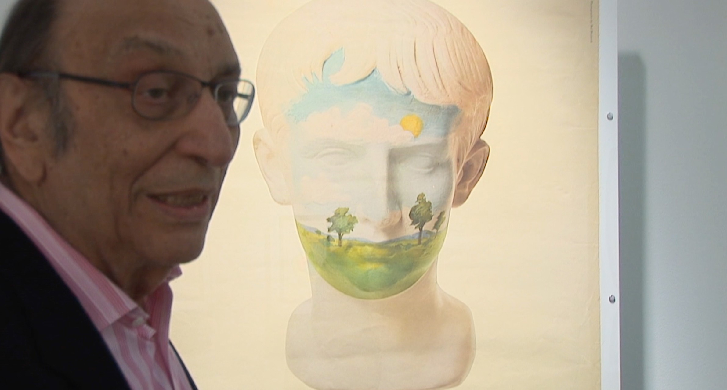 A photo of a man wearing glasses and standing near a white statue, that has it's face painted with a colorful landscape.