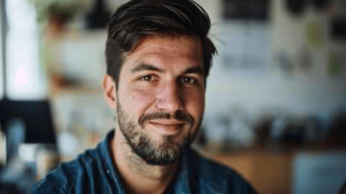 A bearded man with short dark hair smiles warmly in a softly lit workspace.
