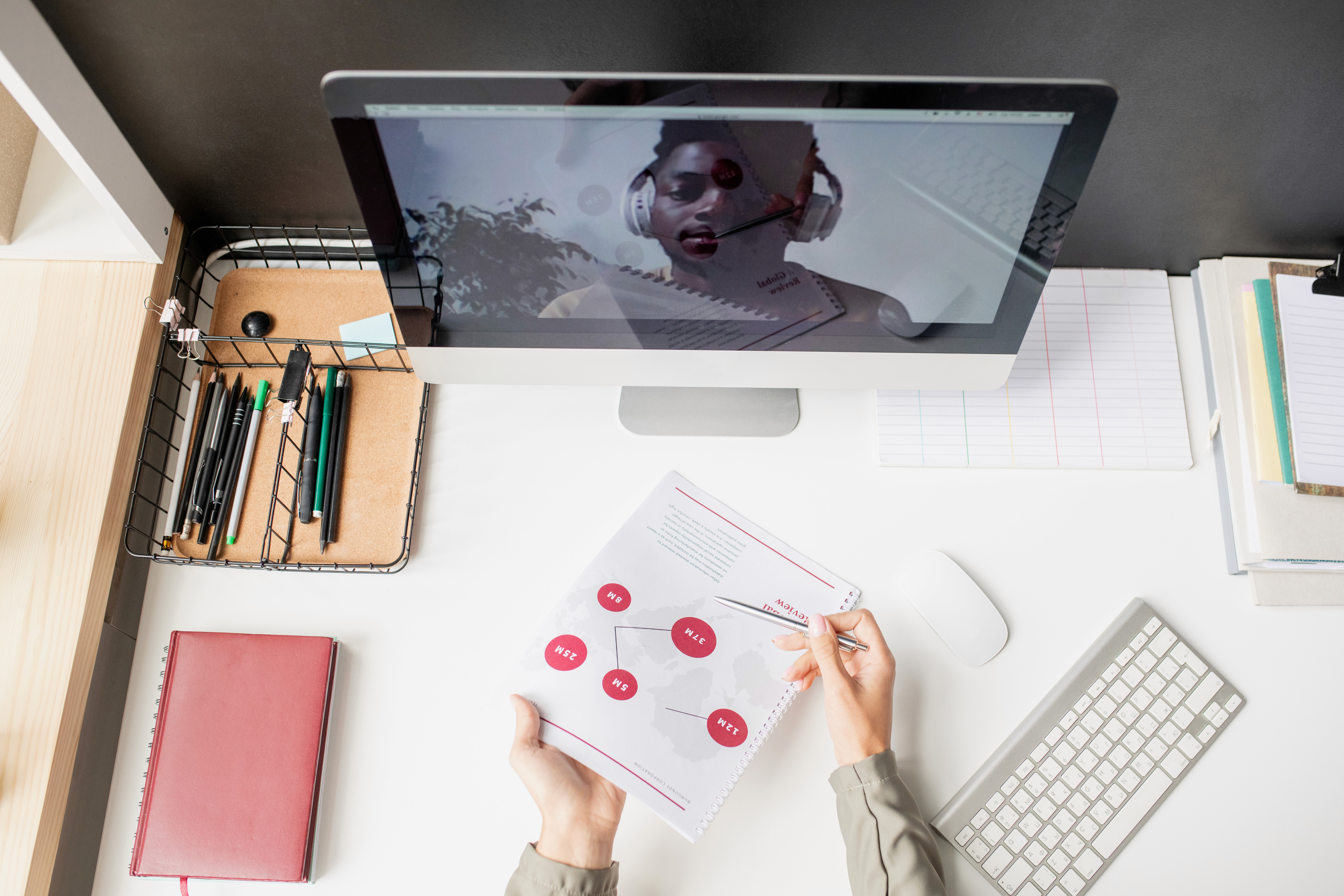 Person holding a pen and analyzing a printed strategy document at a modern desk setup with a desktop monitor, keyboard, and stationery — concept of digital marketing or remote collaboration.