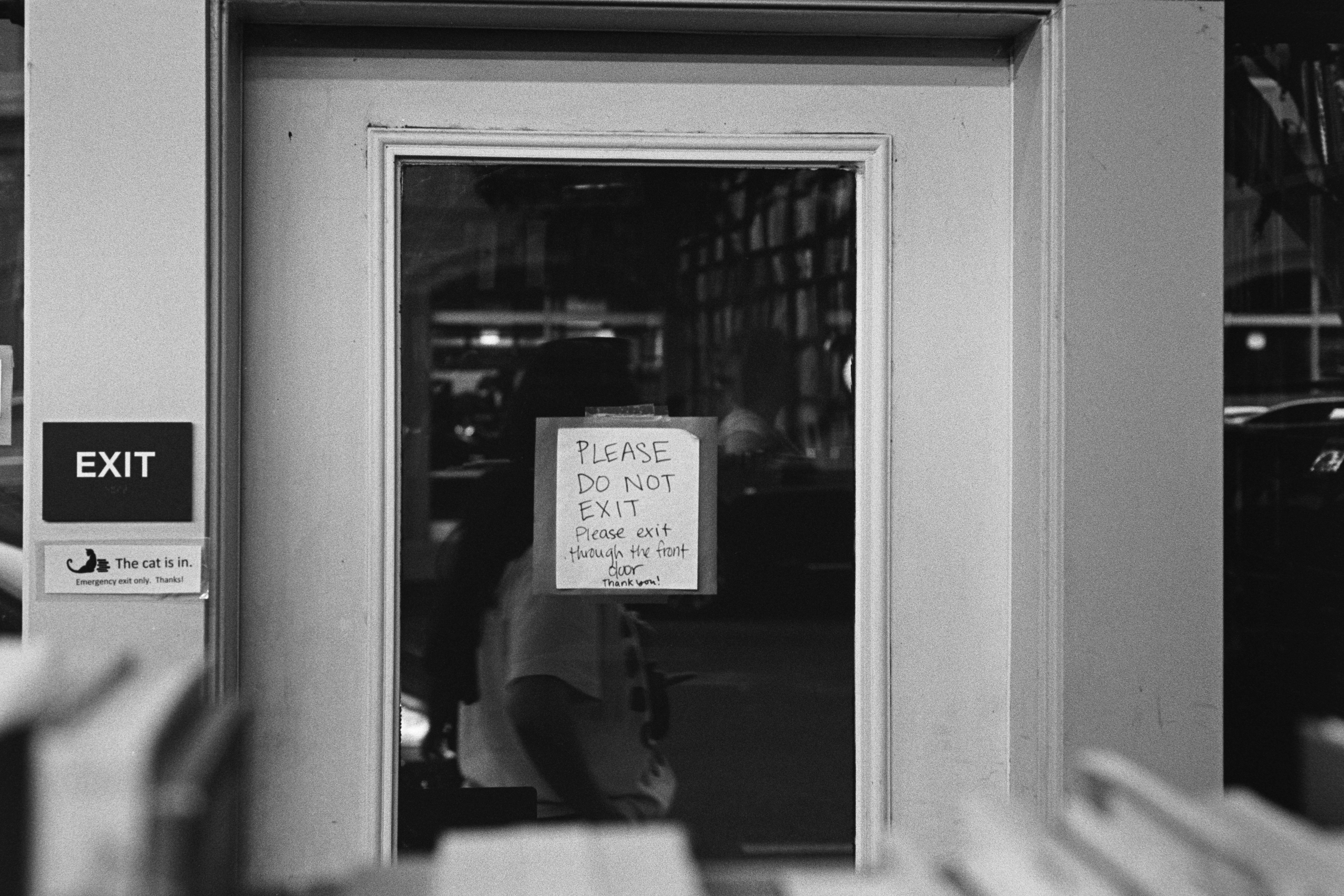A handwritten sign on a glass door asking people not to exit, next to a small sign that says the cat is in.