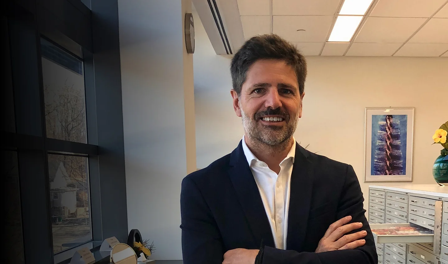A middle-aged man with salt-and-pepper hair and a beard, wearing a white button-down shirt and a dark blazer, stands with his arms crossed and smiles in an office setting.