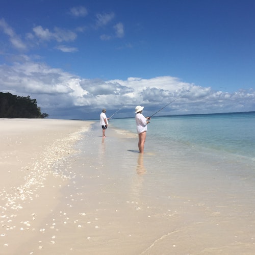 Two people in white hats and clothing fish while standing in shallow, clear water on a wide sandy beach under a partly cloudy sky.