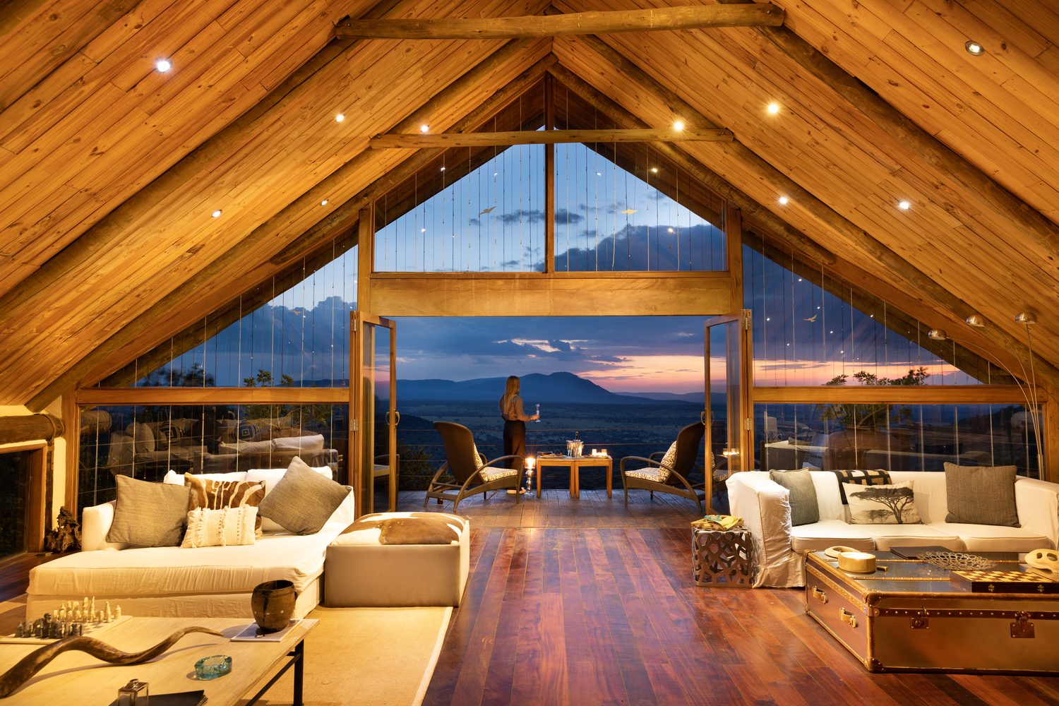 A private villa's vaulted living room at dusk, a guest on the deck beyond