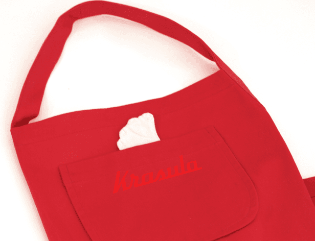 A red apron with a white heart-shaped patch on the chest pocket and the name "Krasula" embroidered in red is displayed against a white background. The apron has a wide neck and a single strap.