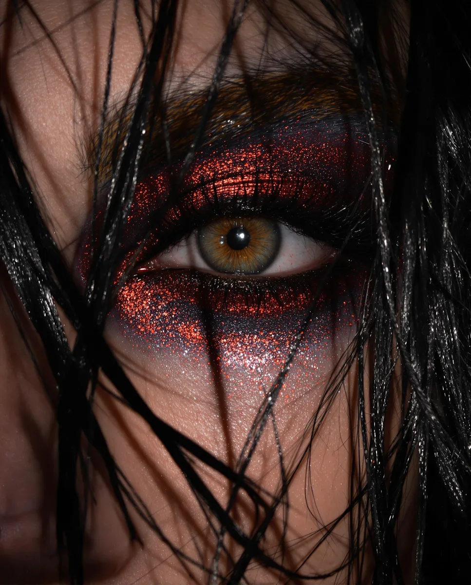 Extreme close-up of a hazel eye with vivid red glitter makeup, partially covered by black strands of hair.