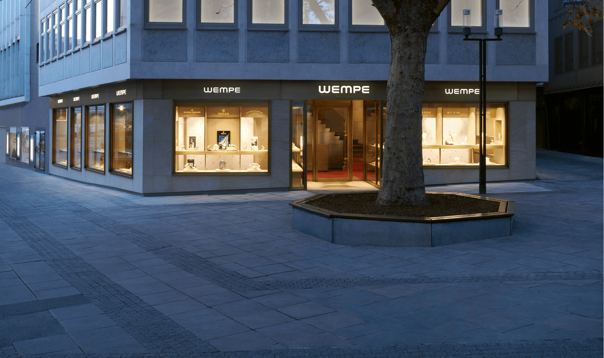 Wempe jewelry store exterior with illuminated displays and a tree in front.  The store is located in a corner building.