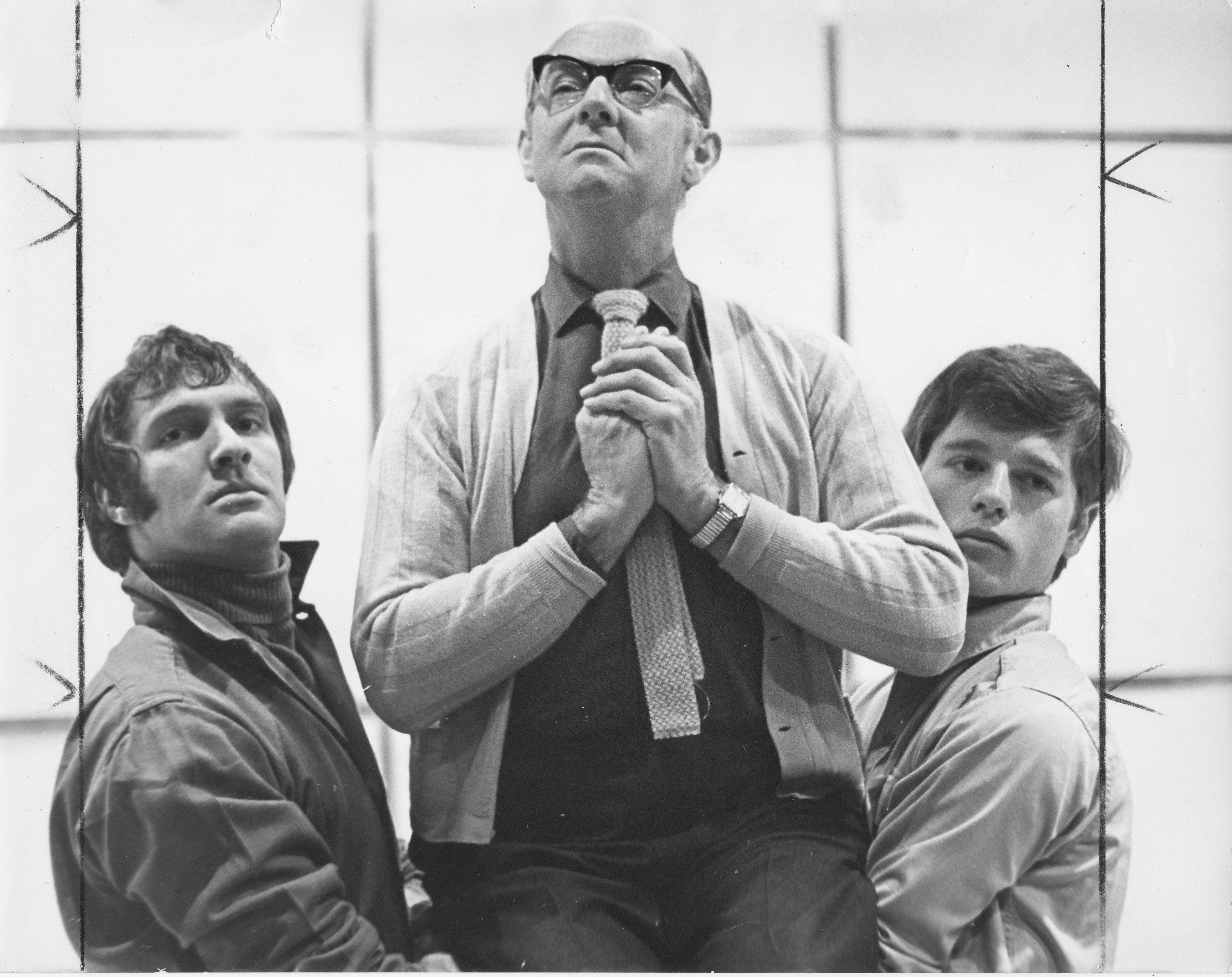 A group portrait of three young adult theatre artists standing and sitting against a dark teal backdrop. A person wearing a navy top and red skirt is seated on a stool in the centre, while two others stand on either side, dressed casually in jeans and T-shirts. All three face the camera with a relaxed, confident expression.