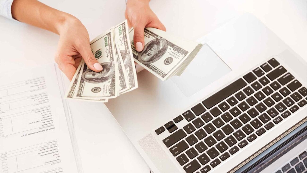 Person counting dollar bills near a laptop on a desk.