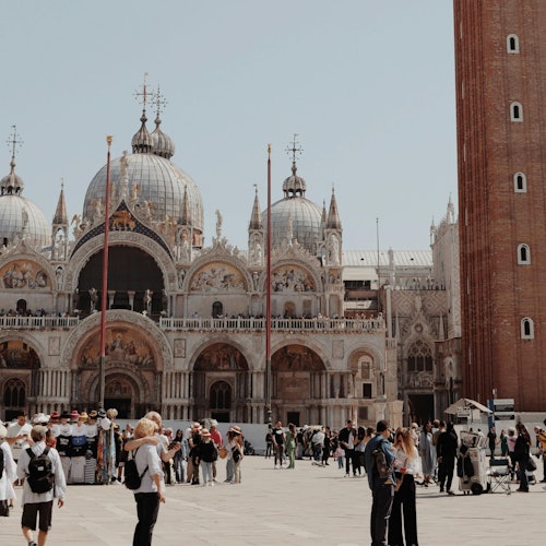 A busy square with people in front of an ornate domed building and a red brick tower under a clear sky.
