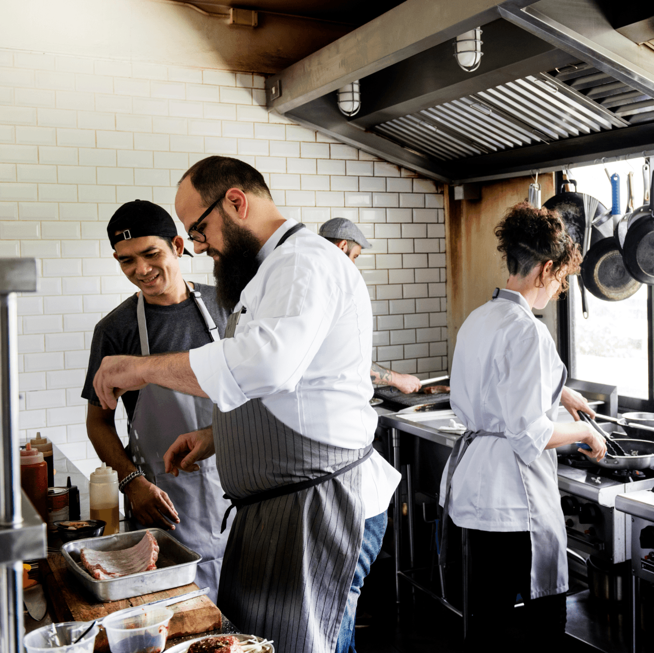Chef avec barbe assaisonnant de la viande sur un plan de travail en bois, entouré de sa brigade dans une cuisine lumineuse aux murs carrelés.