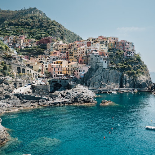 Barevné budovy na skalnatém pobřeží se tyčí nad průzračně modrým mořem s loděmi a bójemi, v pozadí s kopcem.