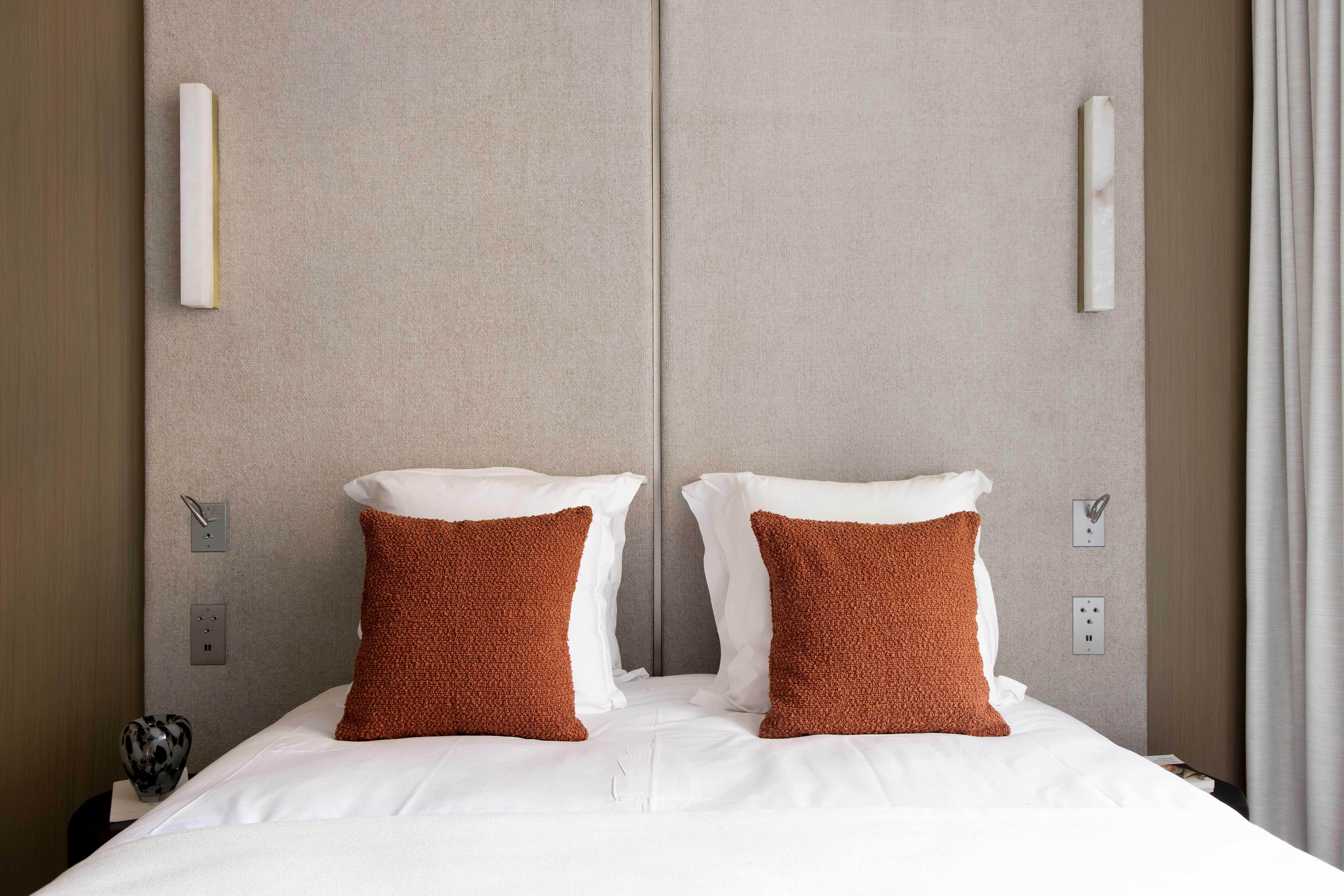 Detail tete de lit beige avec deux coussins terracotta boucles, appliques murales blanches et linge blanc