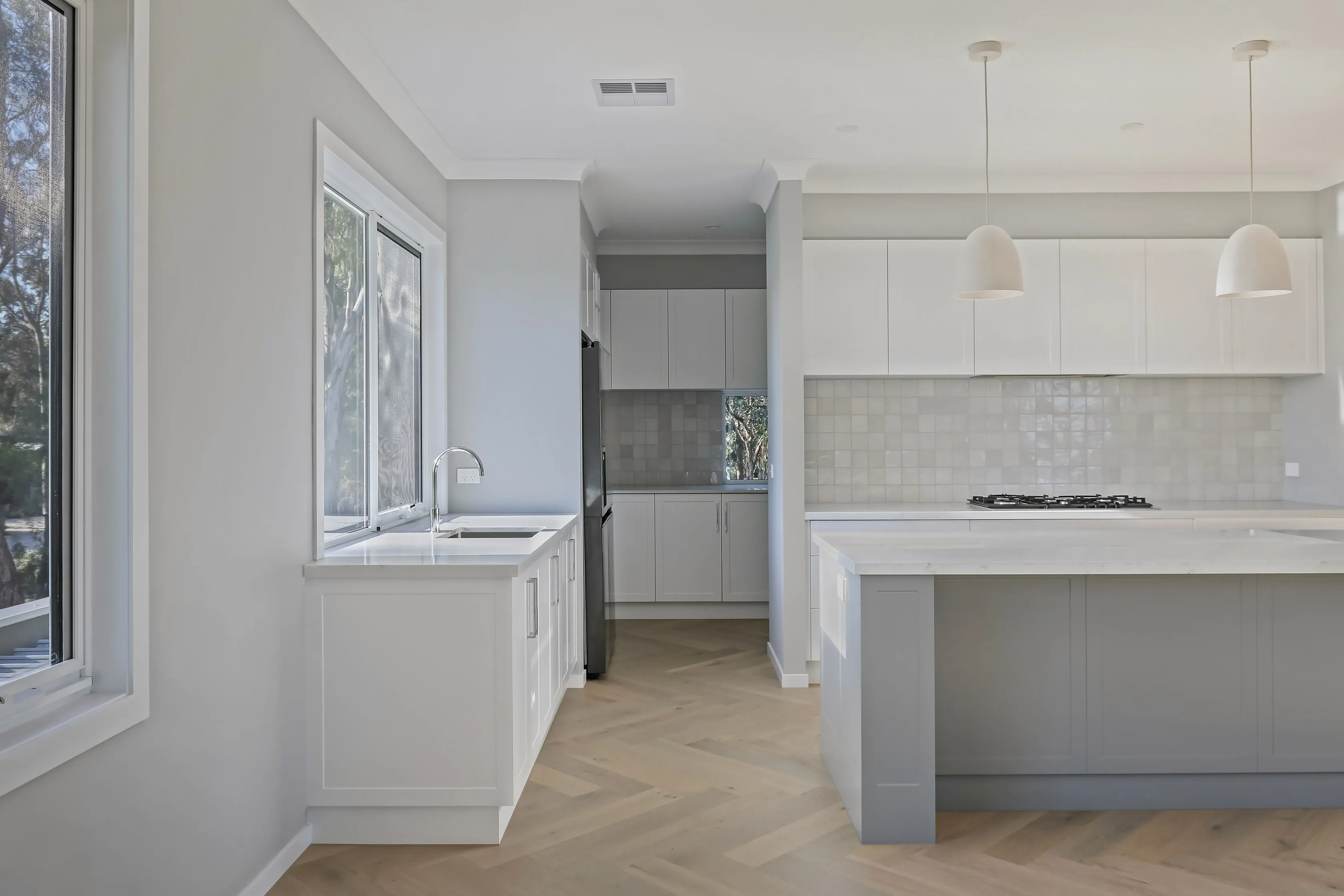 Kitchen and butler's pantry with white shaker cabinetry, a grey island with stone top, white pendants, handmade tile splashback, gas cooktop and a window over the sink to the trees