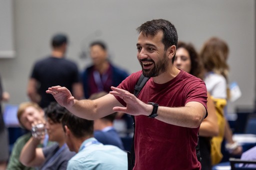a lively moment at an event, featuring a man in a red shirt who appears to be engaging enthusiastically with others around him. The background includes a mix of attendees, creating a vibrant atmosphere. 