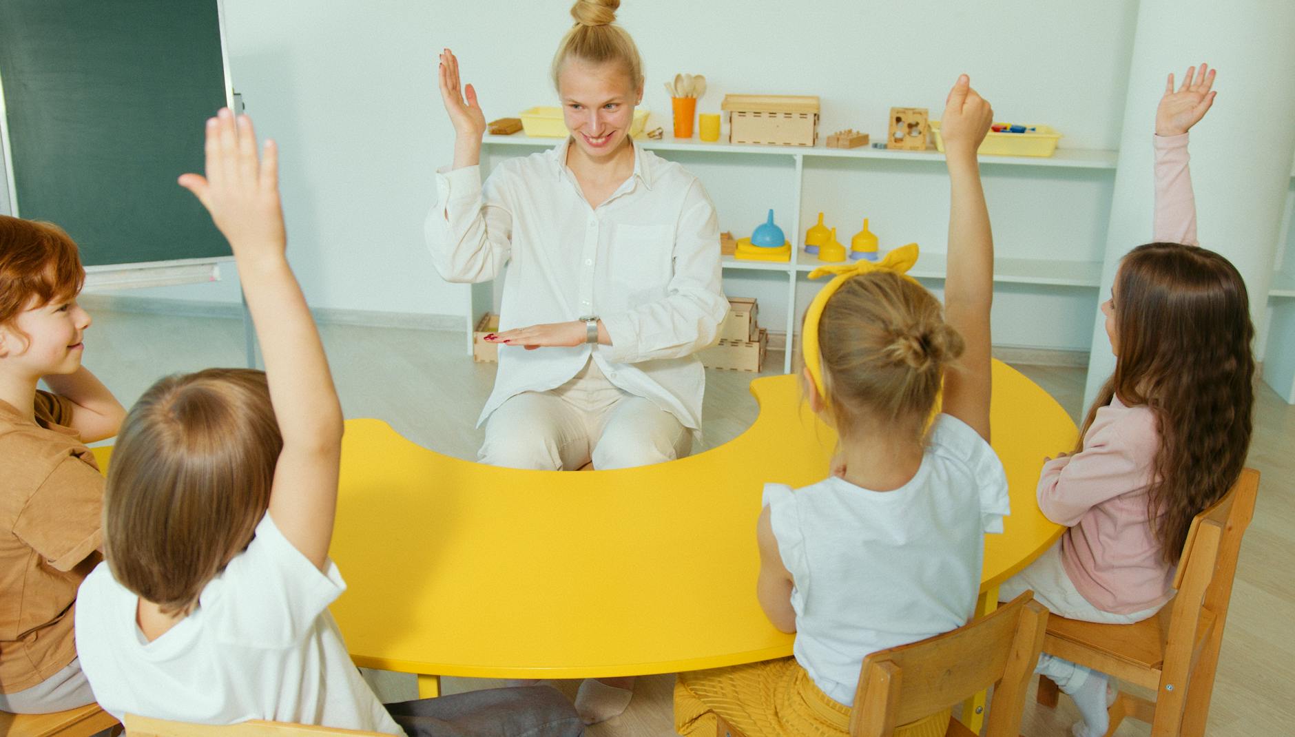 An elementary teacher raising one hand while smiling to signal for silence in a brightly lit classroom.