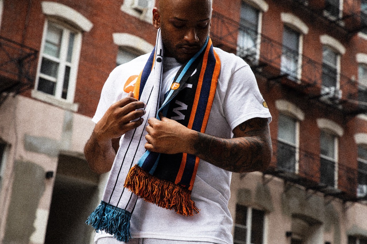 Model adjusting an NYCFC scarf while wearing Staple x NYCFC apparel, photographed outdoors in NYC.