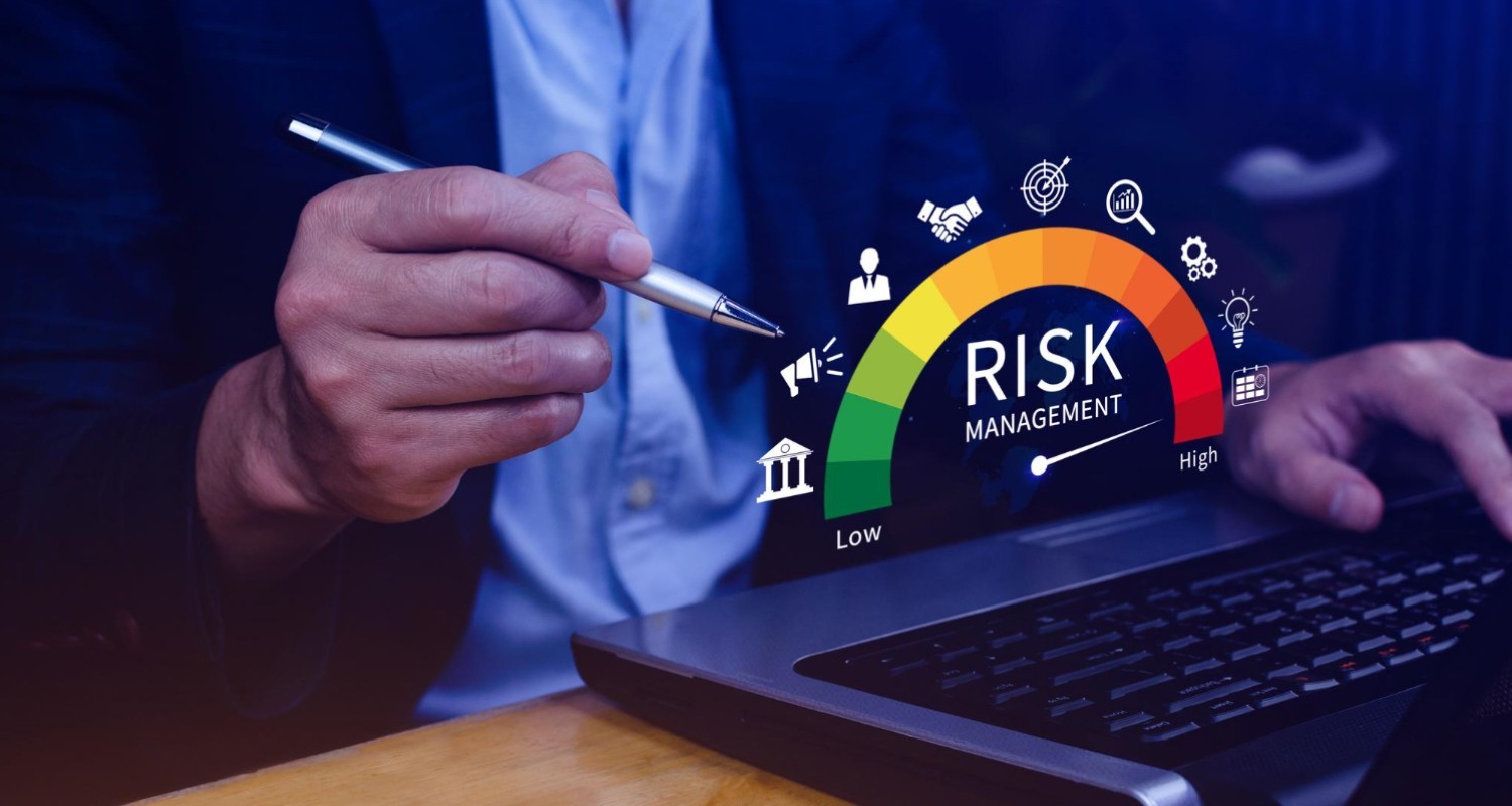 A close-up of a business professional pointing a pen toward a digital risk-management gauge displayed above a laptop, showing a color scale from low to high risk alongside icons representing analytics, banking, users, and financial operations.