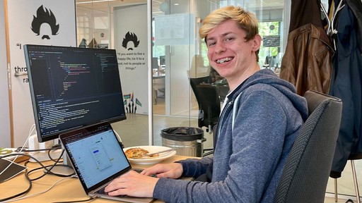 A person is smiling at a desk with a laptop and a monitor in a bright office environment.