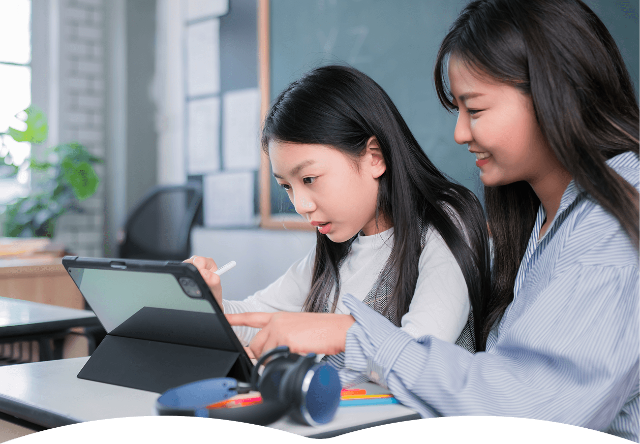 A young student attentively uses a tablet with a digital pen in a classroom setting, guided by an adult, surrounded by educational materials and a chalkboard in the background.