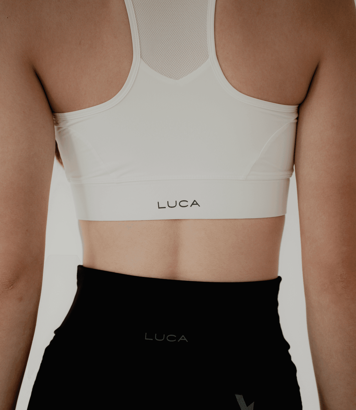 A-close-up-back-view-shot- highlighting-the-design-details-of- the-off-white-sports-bra-and-black- athletic-shorts-The-sports-bra- features-a-racerback-design-with-a- sheer-off-white-mesh-panel-at-the-top- The-"LUCA"-brand-logo-is-centered-in-black-on-both-the- bottom-band-of-the-bra-and-the- shorts-emphasizing-the-matching- activewear-set