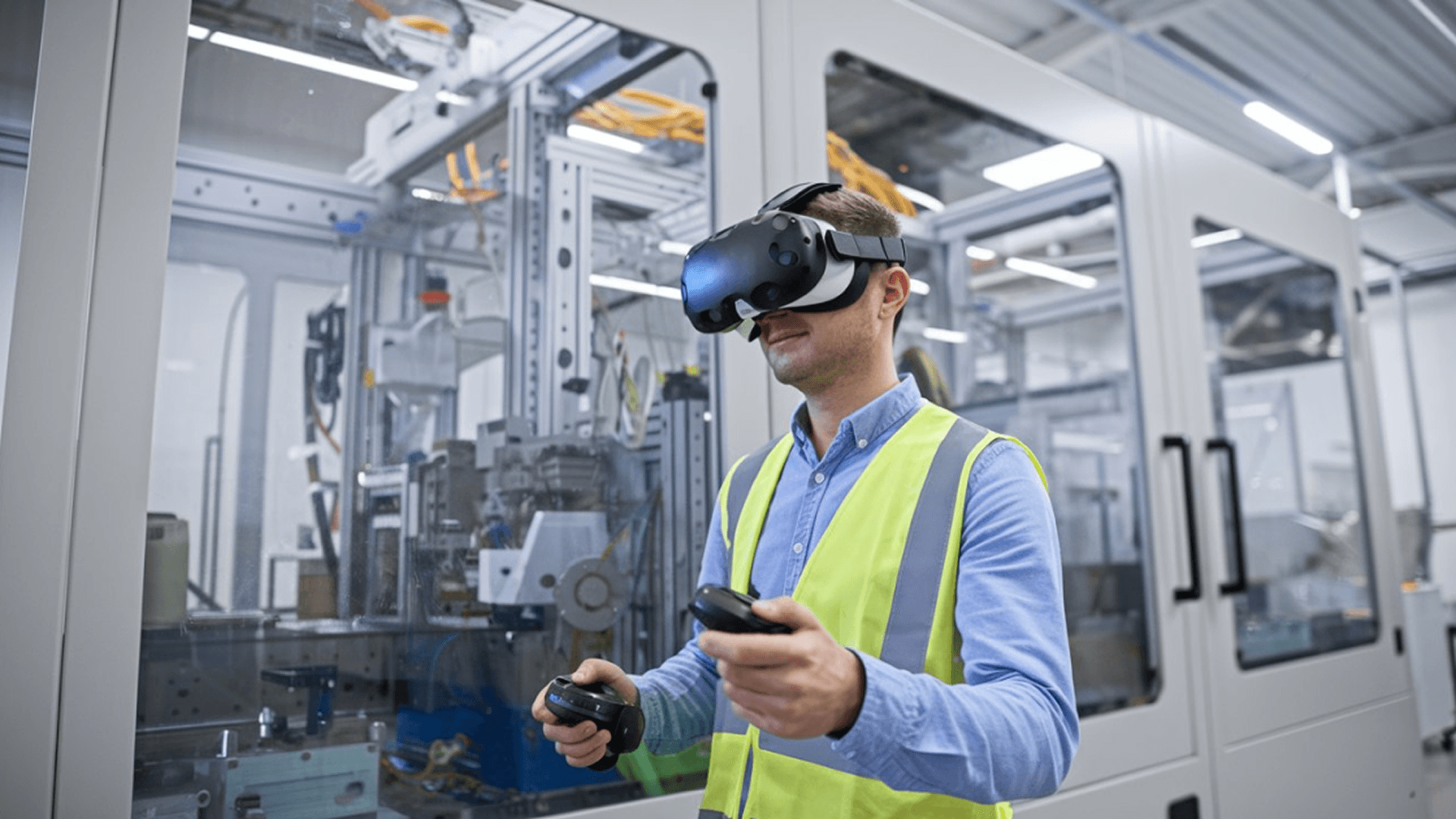A worker wearing a high-visibility safety vest and a virtual reality headset holding controllers, participating in an immersive training simulation inside a modern industrial manufacturing facility.