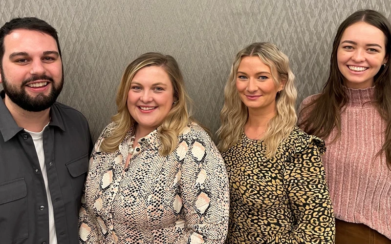 Four people standing together in front of a textured beige wall, wearing patterned and solid-colored tops including snake print, leopard print, and a pink ribbed sweater.
