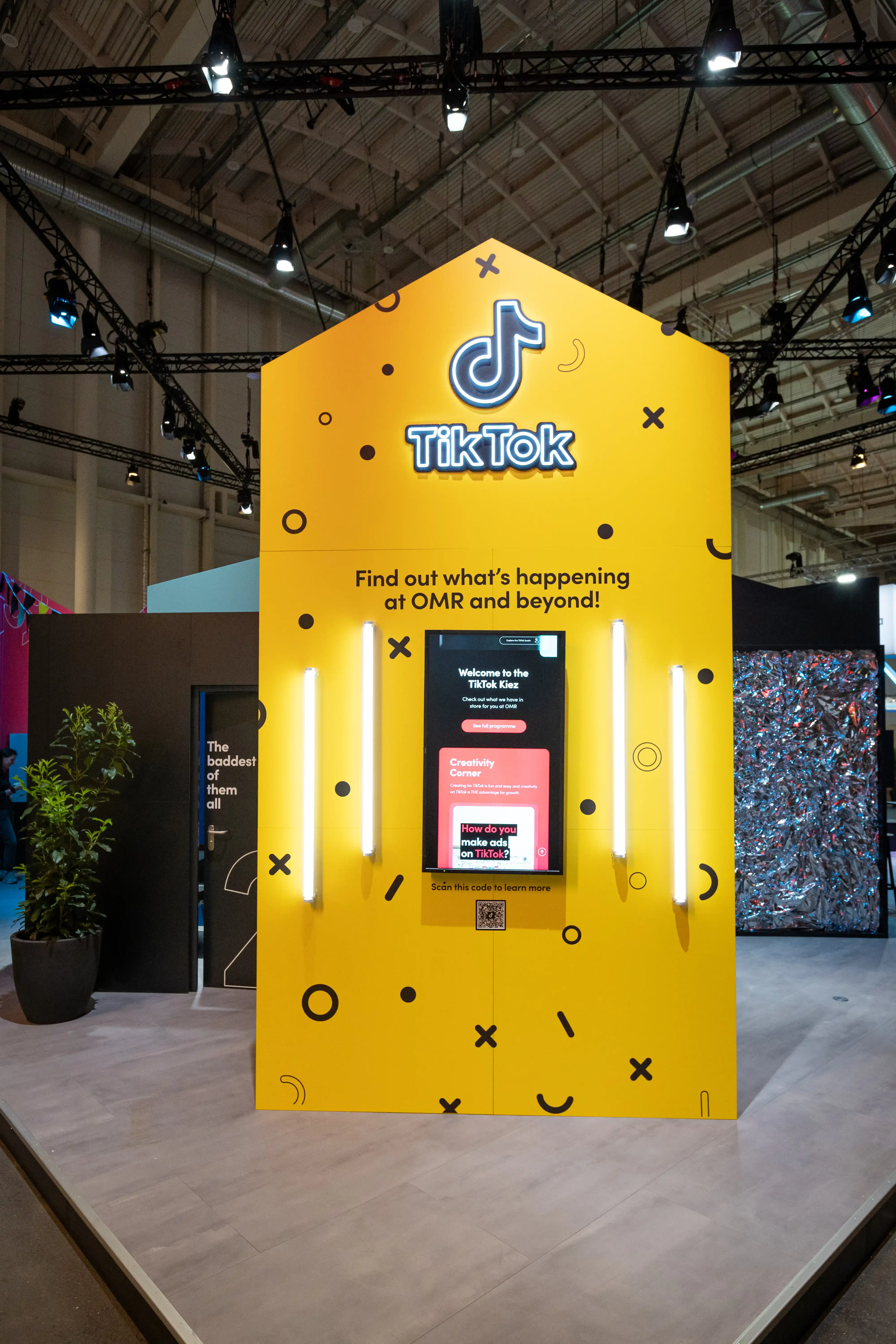 Yellow house-shaped facade with TikTok logo, neon tubes and interactive kiosk screen at the TikTok booth at OMR.
