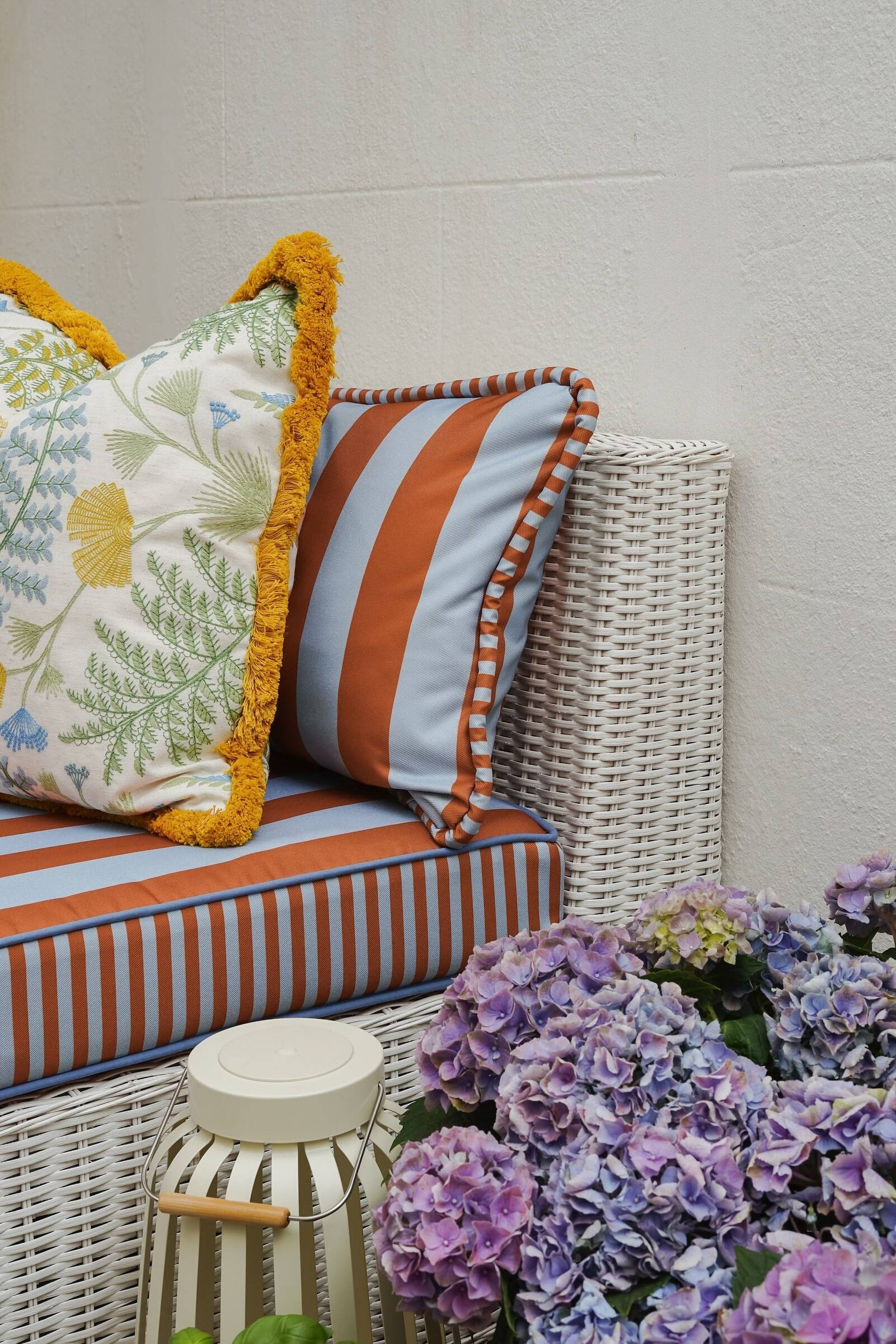A white wicker sofa with colorful striped and pattern matched embroidered pillows, a small white side table, and purple hydrangea flowers in front against a beige wall.