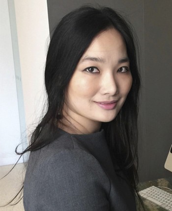 A woman with long black hair smiles gently while sitting against a neutral-colored wall.