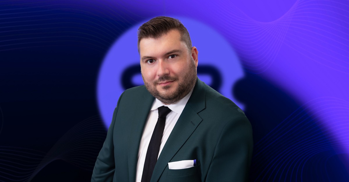 Wiktor Garczyński, CPO of Sb Software, with dark hair and a beard, wearing a dark green blazer, white shirt, and black tie, smiling at the camera. The blurred background features the "SB Software" logo.