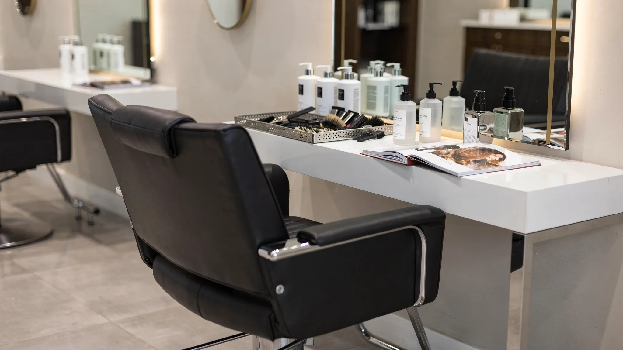 A clean luxury salon station with styling chair, mirror, hair products, combs, clips, and a client inspiration photo booklet on the counter.