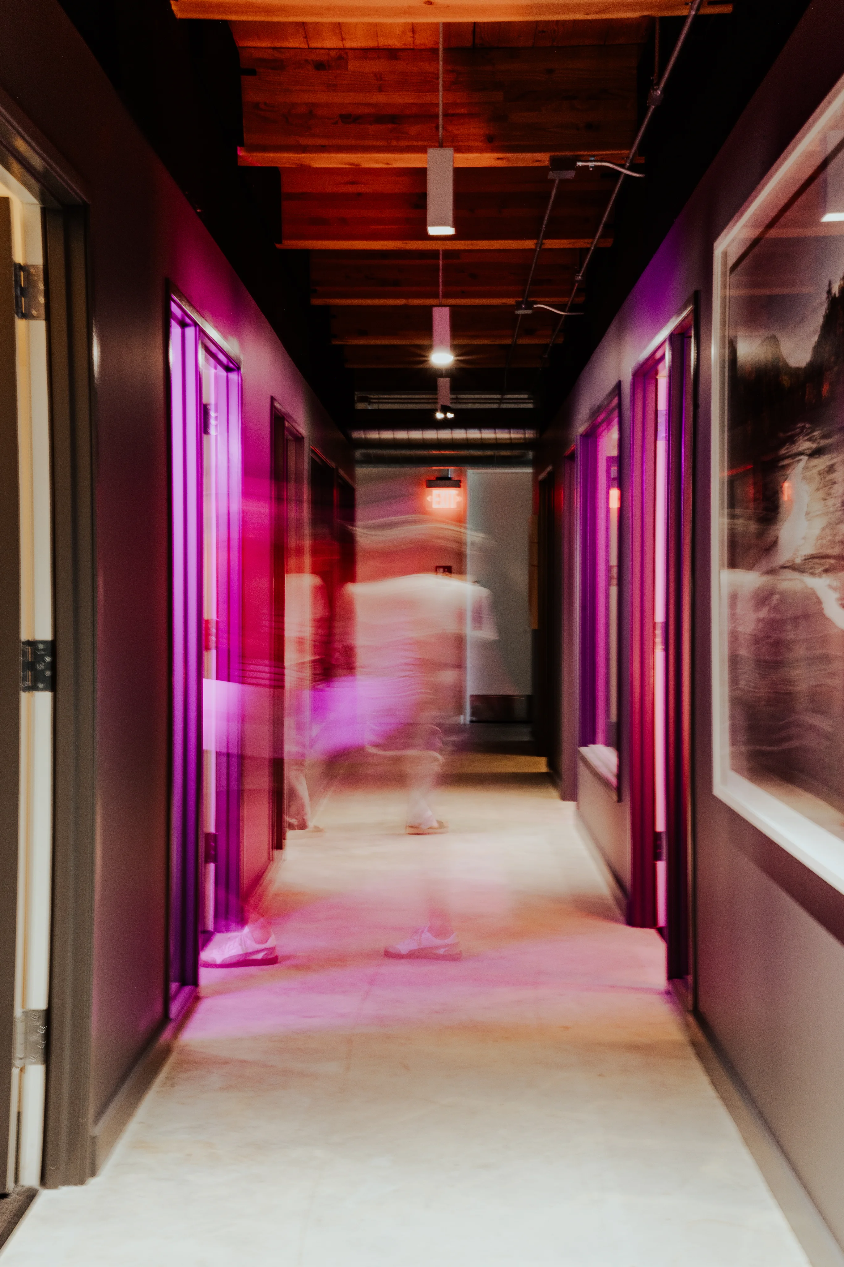 A motion-blurred figure walking through a neon pink-lit office corridor with exposed wood ceiling beams and framed artwork on the walls