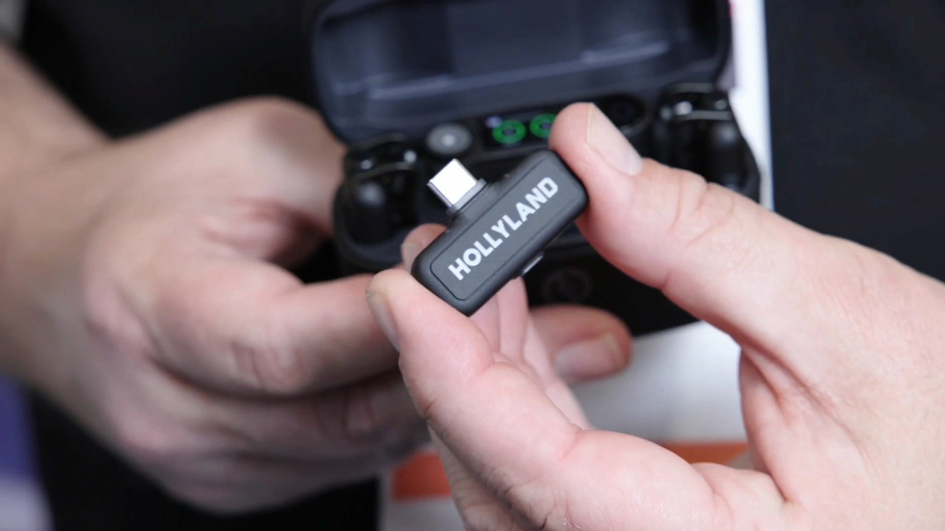 Hands holding a Hollyland wireless microphone receiver plug next to its open charging case.