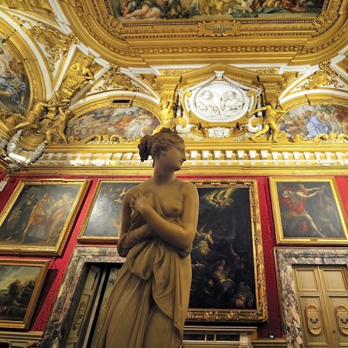 A stone statue of a woman stands in an ornate room with gold details, large framed paintings, and a richly decorated ceiling.