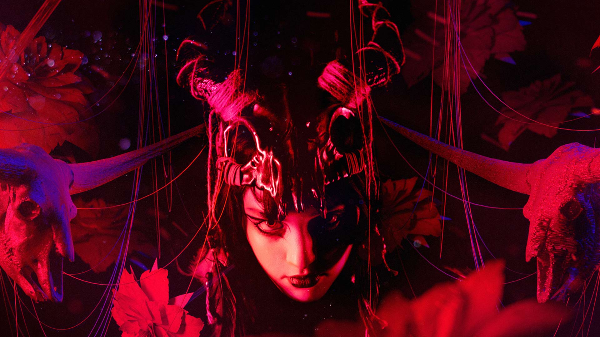 A woman with dark hair and elaborate headdress adorned with horns and ribbons stands centrally in the image. Her face is partially obscured by the headdress, and she gazes directly at the viewer. The background is dark with red and purple lighting effects, and stylized floral patterns are visible. Horn-like structures extend outward from either side of her head. The lighting creates a dramatic, theatrical atmosphere.