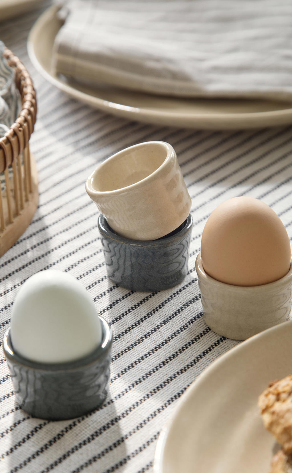 Trois coquetiers en grès aux tons gris et beige avec des œufs, posés sur une nappe à rayures fines, avec plateau et panier en osier