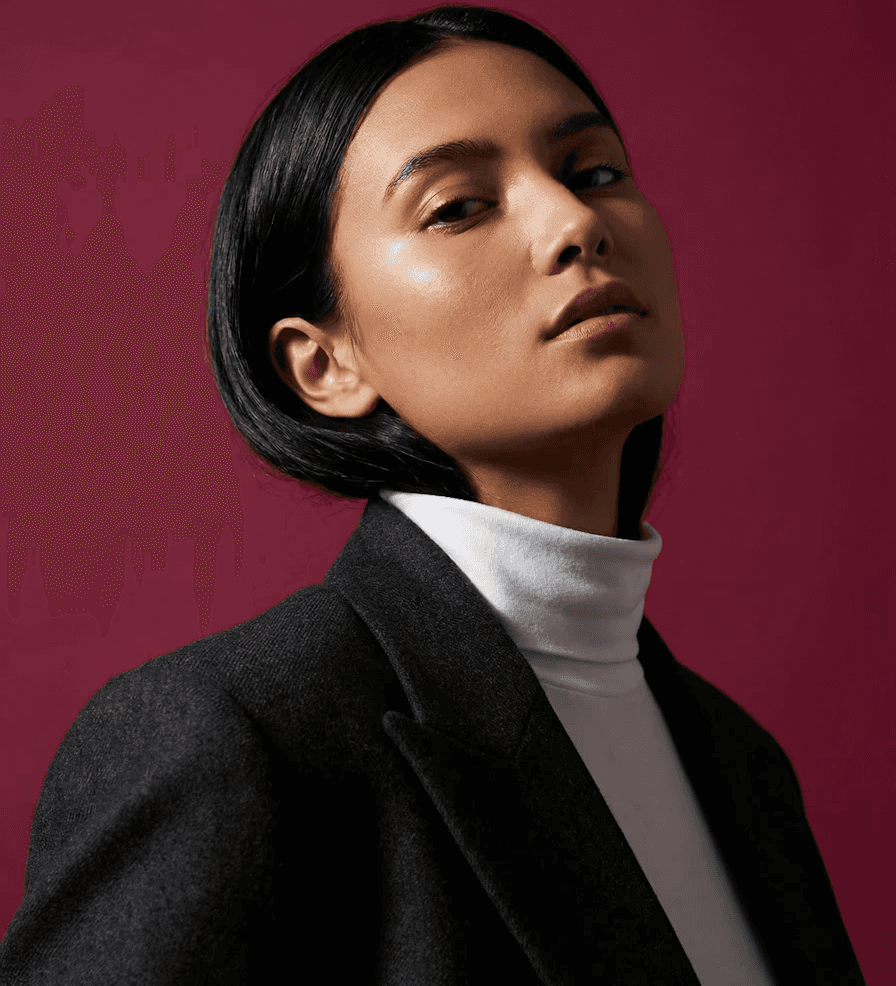 Elegant editorial-style profile shot of a woman with straight black bob and glowing dewy skin, gazing upward; she wears a tailored charcoal blazer layered over a crisp white turtleneck, rich burgundy background creating dramatic contrast and highlighting her poised expression.
