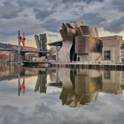 Modern building with metallic, abstract architecture reflected in a calm river, with a red and green bridge in the background.