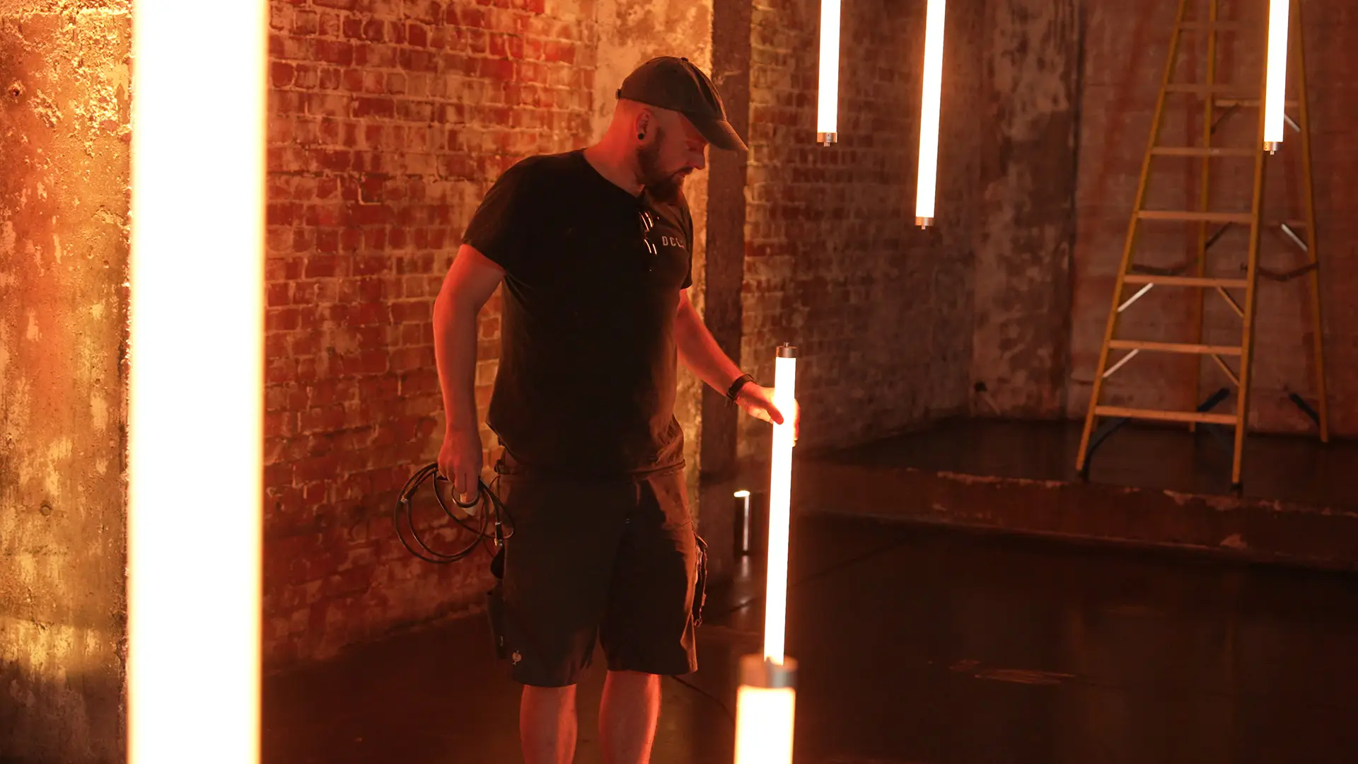 Lighting technician positioning vertical LED tube lights inside an industrial warehouse studio for the Wired x Kyndryl broadcast.