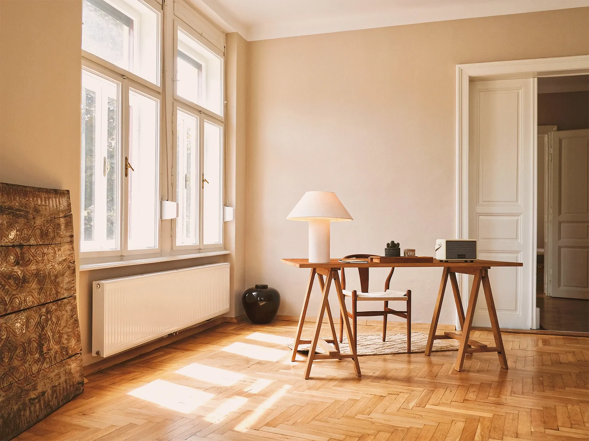 Sunlit apartment interior with herringbone parquet floor, tall windows and a wooden desk with a white lamp