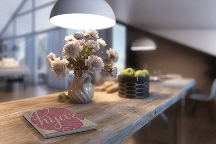 A light wood table holds a vase of cream-colored flowers, a dark blue container with green apples, and a pink book with the word "hiya" written on it. A large white pendant light hangs above the table. The background is out of focus, showing a window with curtains and a portion of a dining area.