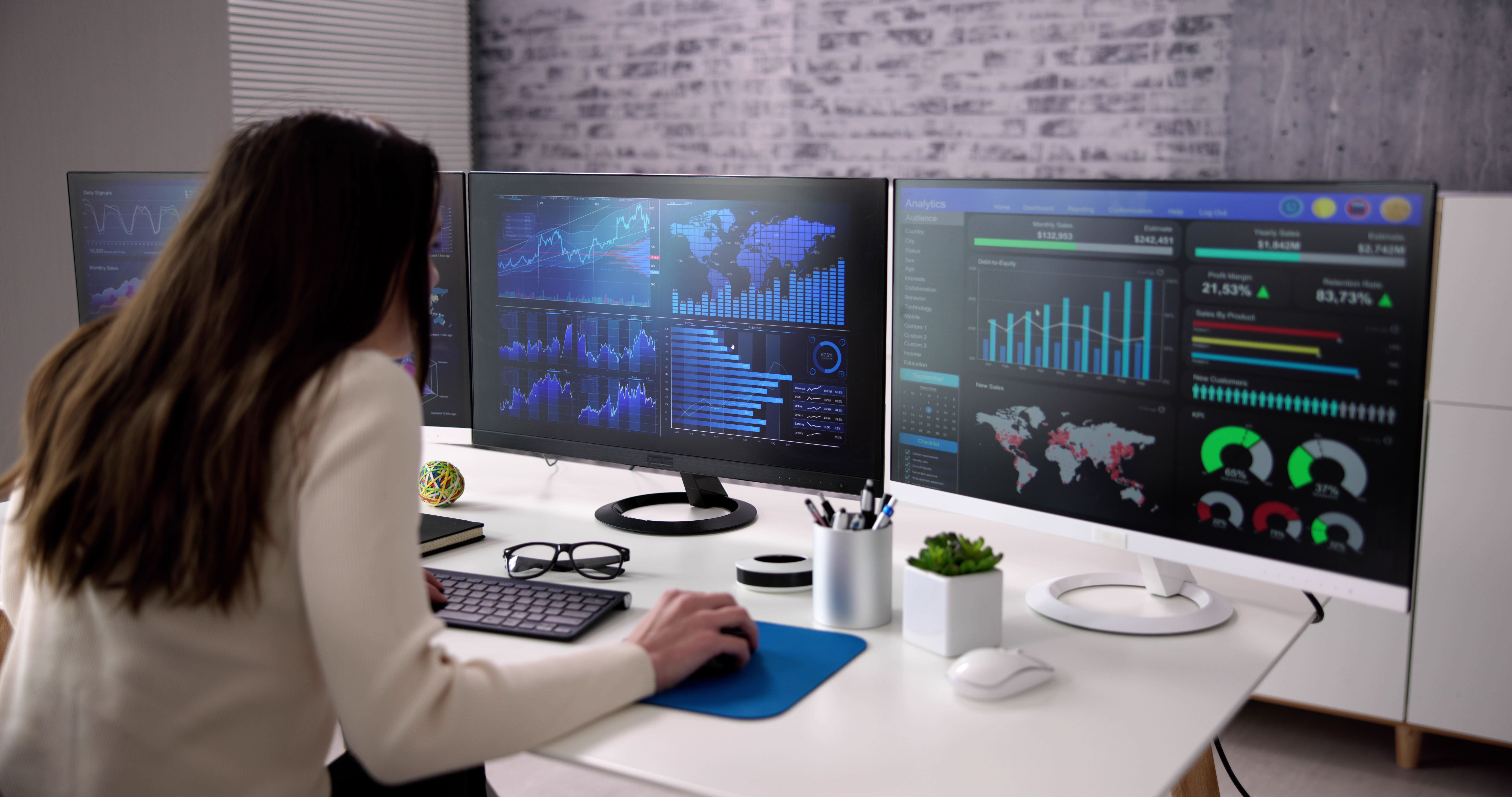 A person sits at a modern desk, analyzing data on multiple computer monitors displaying various graphs and charts, highlighting trends and statistics in a professional office setting.