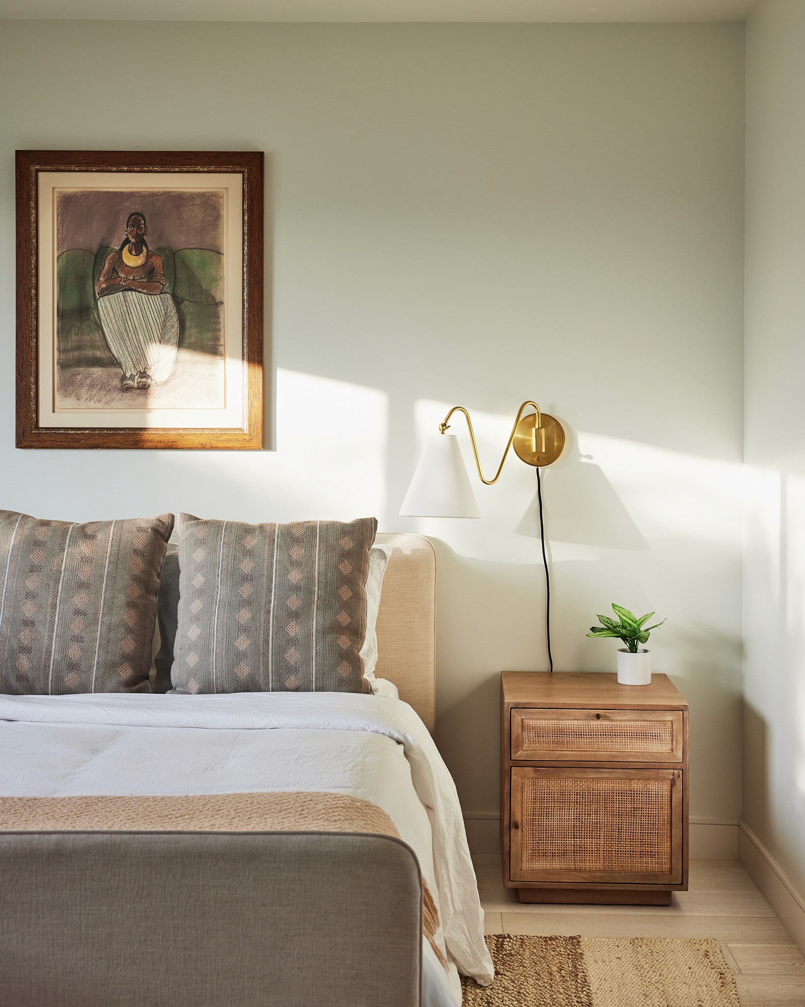 Guest bedroom with natural tones and textures, wall mounted reading light, upholstered bed, wood side table, grey accent pillows and blanket with sunlight pouring through the side window by Jubilee Interiors in Los Angeles, California
