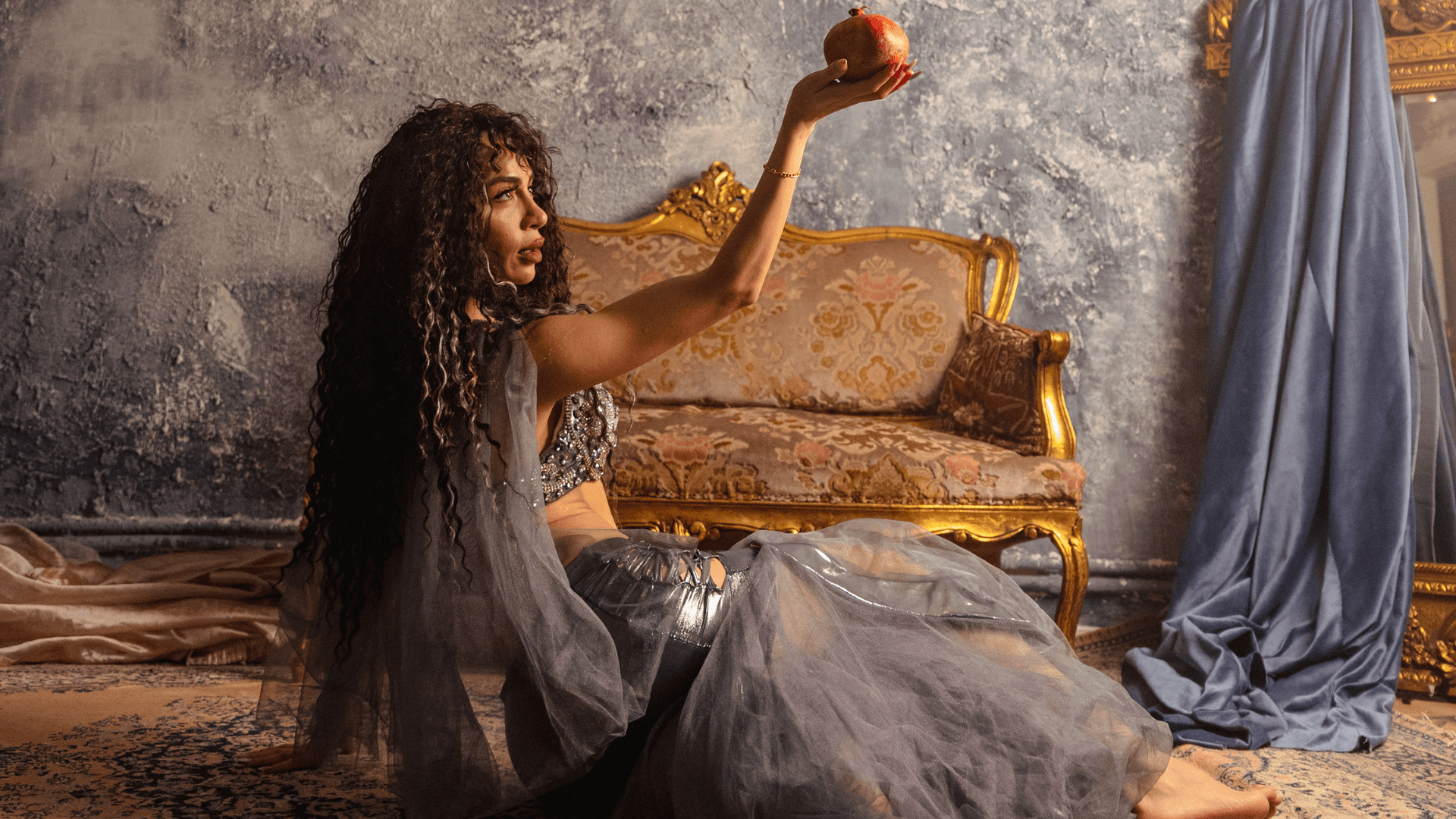 A belly dancer in a silver costume sits gracefully on the floor, holding a pomegranate aloft in a symbolic pose, with an ornate vintage sofa and textured wall in the background.