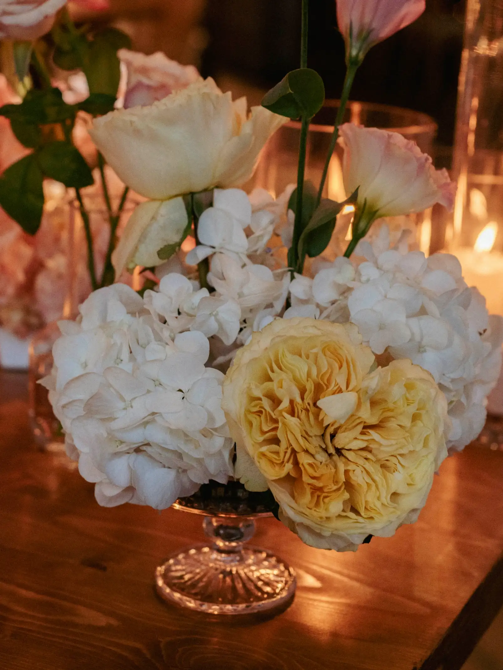Arreglo floral en copa de vidrio con rosas, hortensias y flores amarillas, iluminado por la luz de las velas.