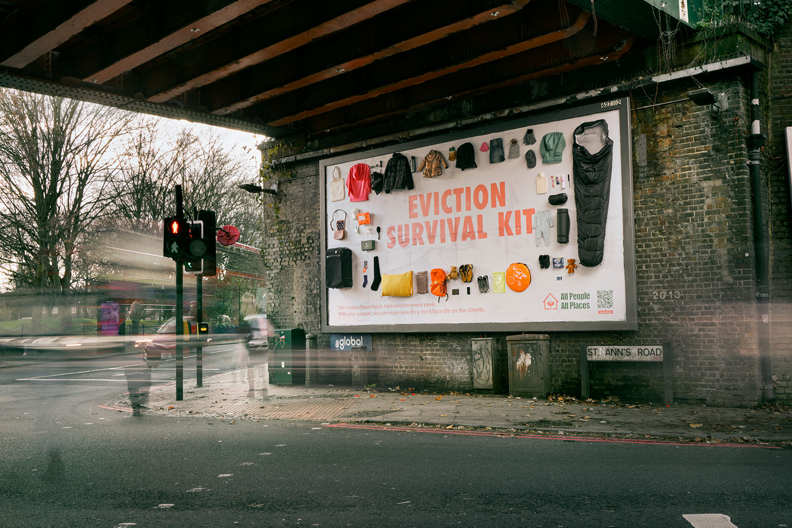 A billboard with clothes and sleeping bags underneath a bridge. The billboard says Eviction Survival Kit