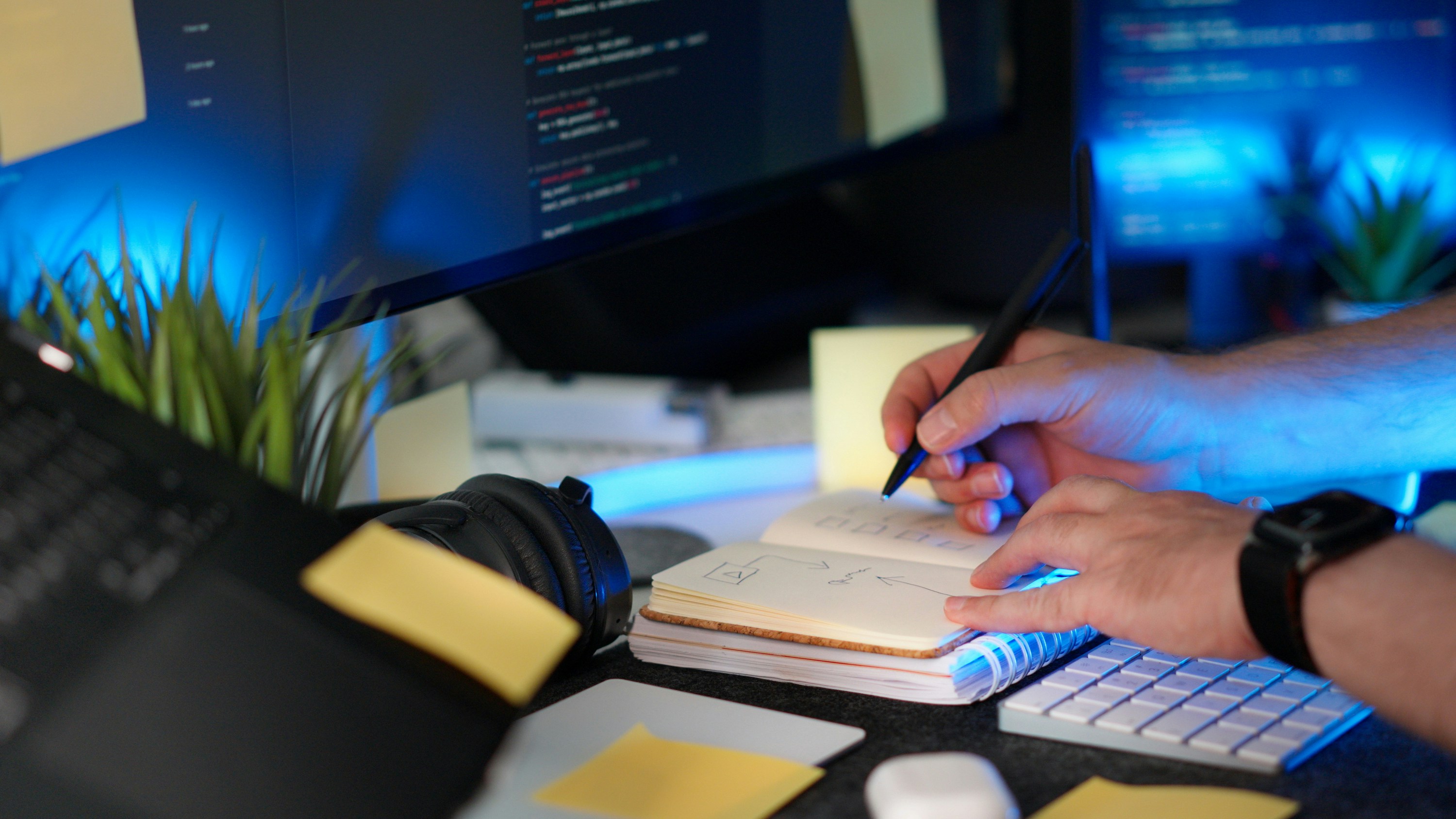 Person writing notes at a computer workstation with code on the screen, keyboard, headphones, and sticky notes on the desk.