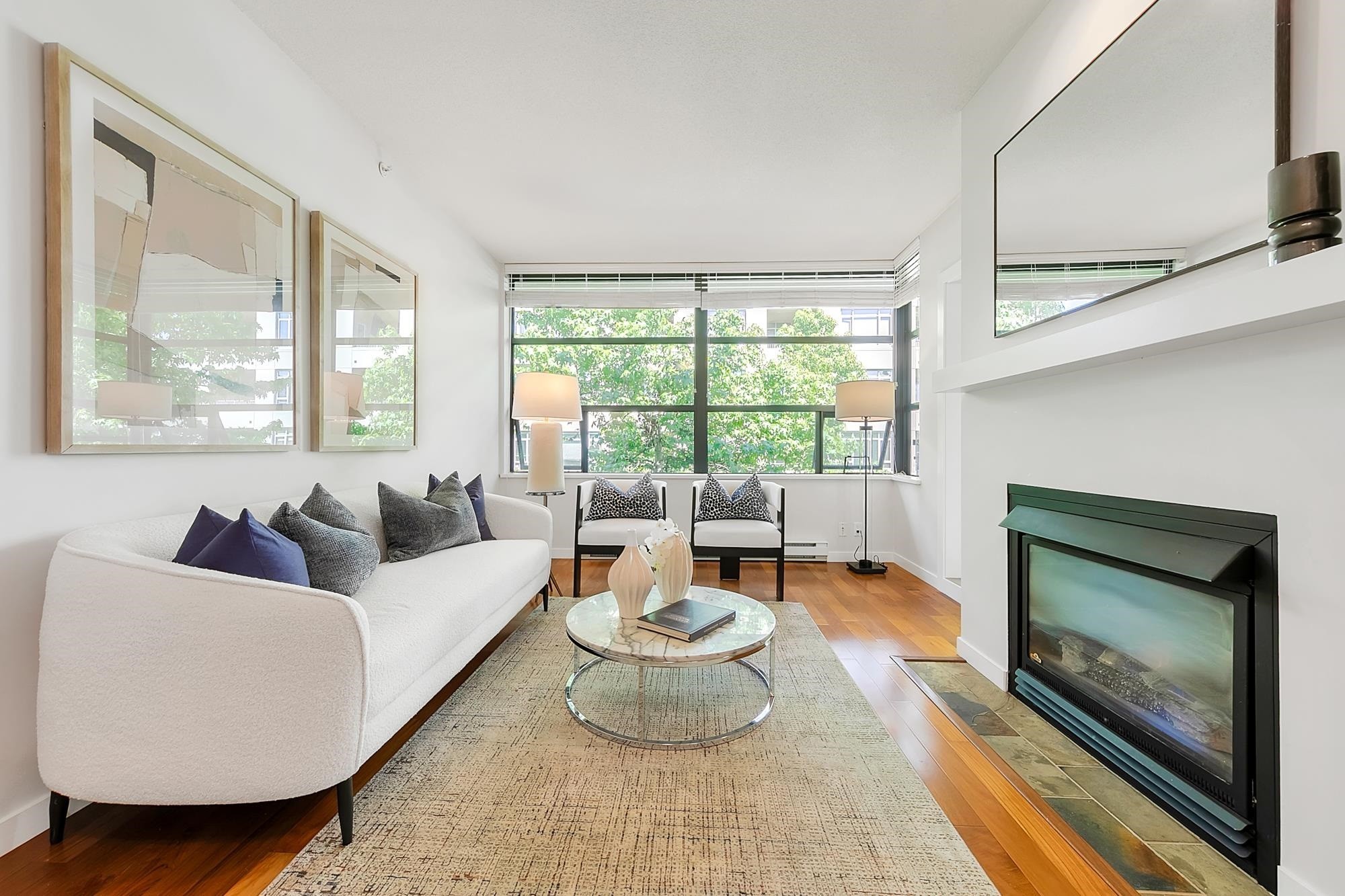 A bright, modern living room with a white sofa, a coffee table, and a fireplace, featuring large windows.
