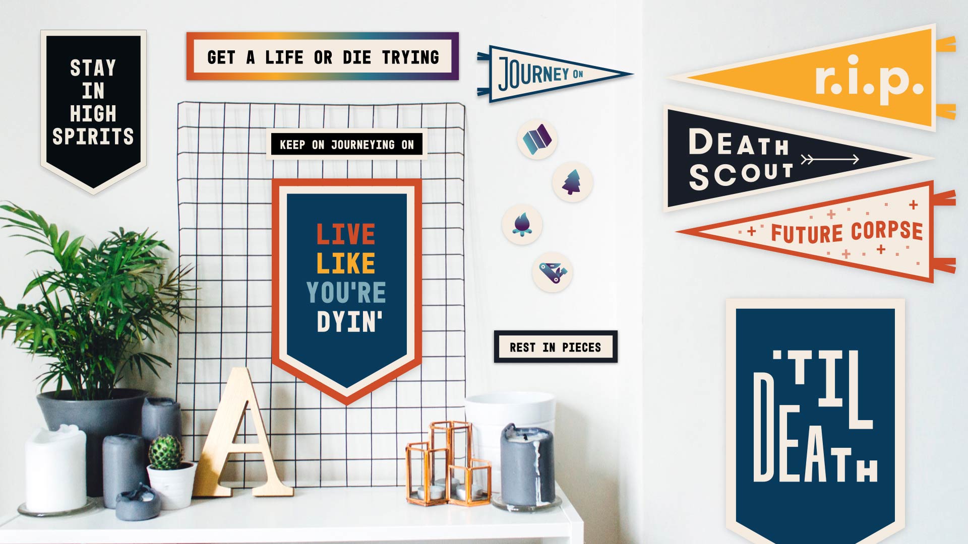 A collection of pennants and wall decorations on a white wall. The pennants display various messages, including “STAY IN HIGH SPIRITS,” “GET A LIFE OR DIE TRYING,” “JOURNEY ON,” “DEATH SCOUT,” “FUTURE CORPSE,” and “REST IN PIECES.” A white grid-patterned board holds “LIVE LIKE YOU’RE DYIN’!” and “KEEP ON JOURNEYING ON.” A potted plant, candles, a wooden “A,” and decorative metal objects are arranged on a white surface.
