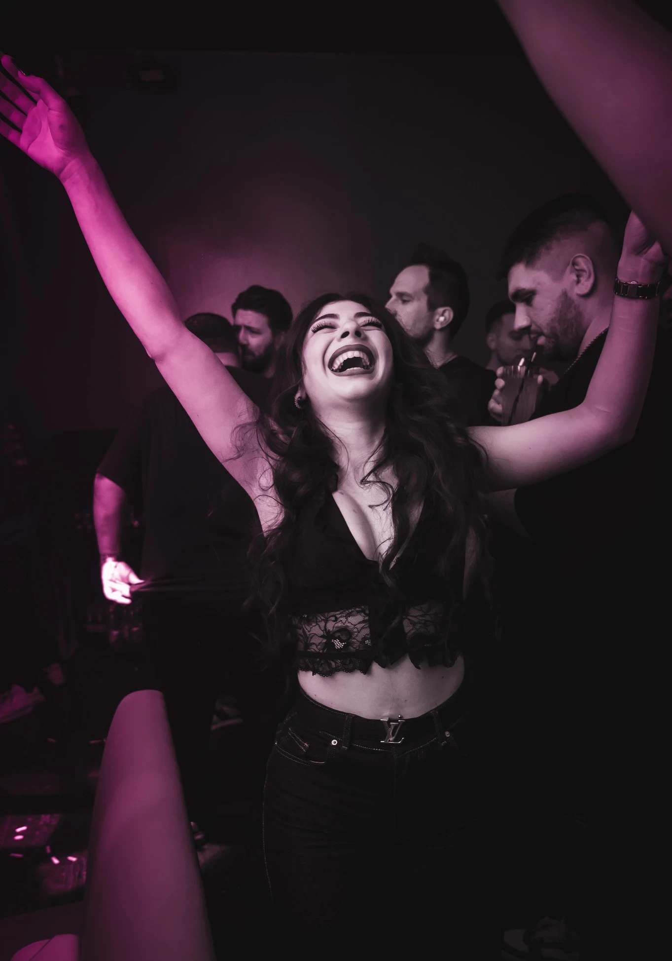 A person with long dark hair smiles and dances in a crowd at the RHB party, their arms raised. They are wearing a black strapless top and an "RHB" necklace.